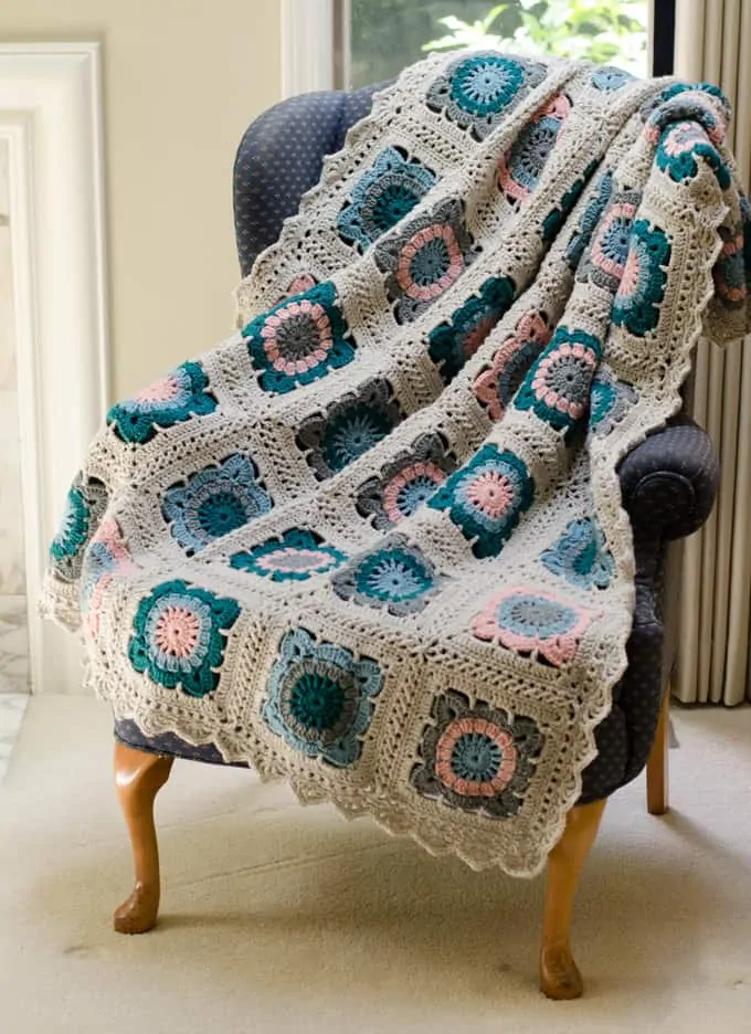 Crochet afghan in blue, teal, pink , gray and off white yarn on a chair in front of a window