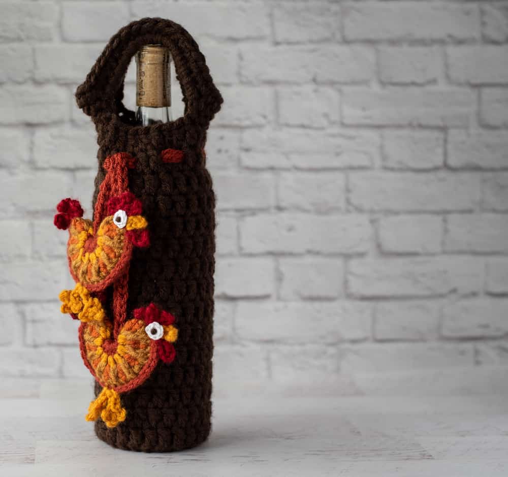 crochet rooster appliques hanging from brown wine bottle cover with bottle of wine