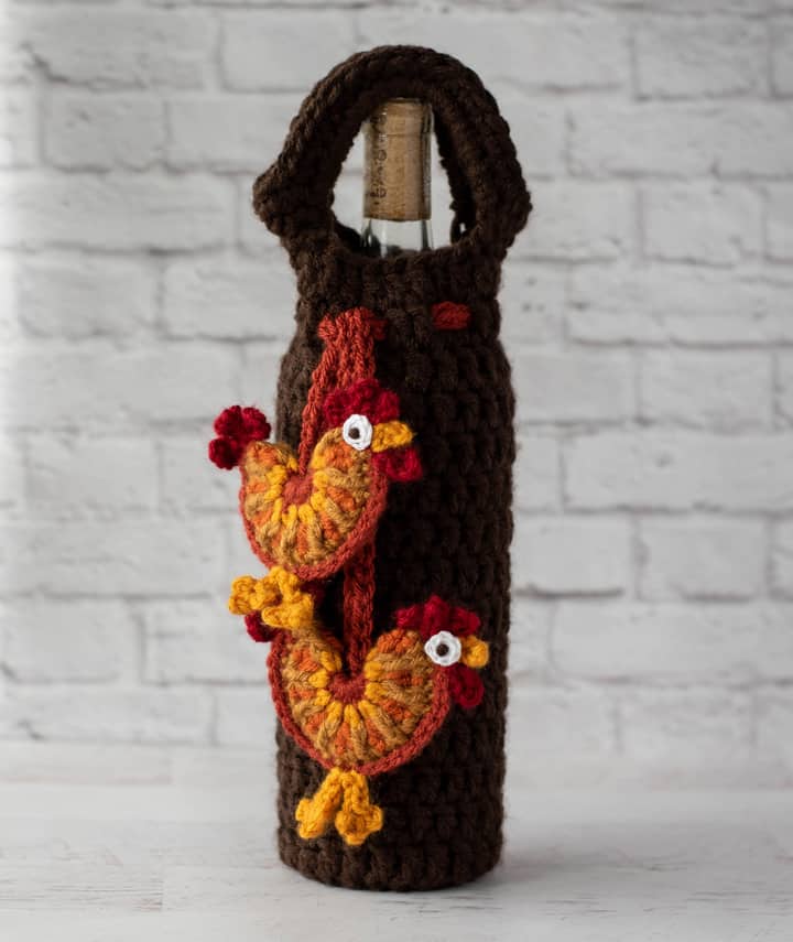 crochet rooster appliques hanging from brown wine bottle cover with bottle of wine