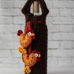 crochet rooster appliques hanging from brown wine bottle cover with bottle of wine
