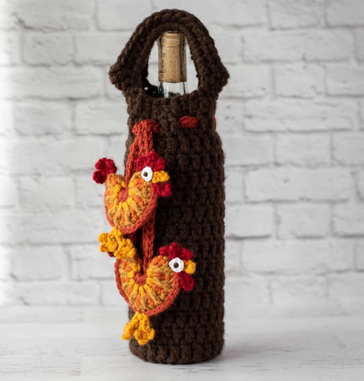 crochet rooster appliques hanging from brown wine bottle cover with bottle of wine