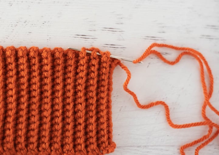 yarn needle sewing top of orange crochet ribbed fabric