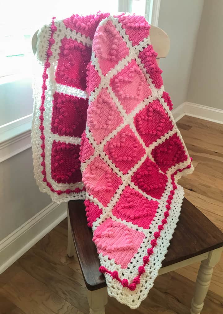crochet afghan with pink heart squares and a white lacy like border displayed on a chair by a window