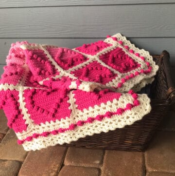 crochet afghan with pink heart squares and a white lacy like border in a basket on a porch