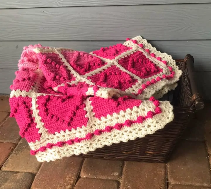 crochet afghan with pink heart squares and a white lacy like border in a basket on a porch