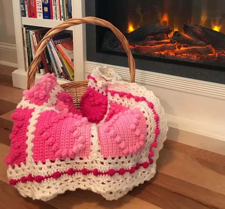 crochet afghan with pink heart squares and a white lacy like border by a fireplace and bookcase