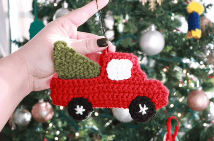 hand holding red crochet truck carrying tree ornament in front of a christmas tree