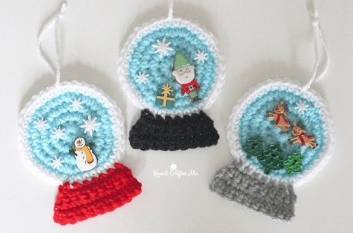 three crochet flat snow globe ornaments: one with a snowman and red bottom, one with an elf, gift, and black bottom, and one with two reindeer, three trees, and a grey bottom