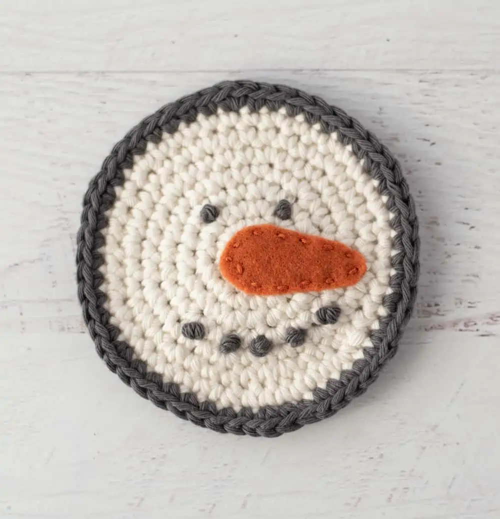 cream and gray crochet snowman coaster with orange carrot nose