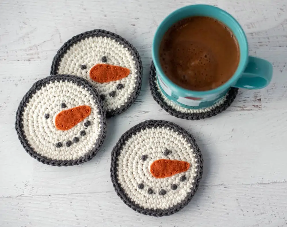 cream and gray crochet snowman coasters with orange carrot nose with cup of hot cocoa