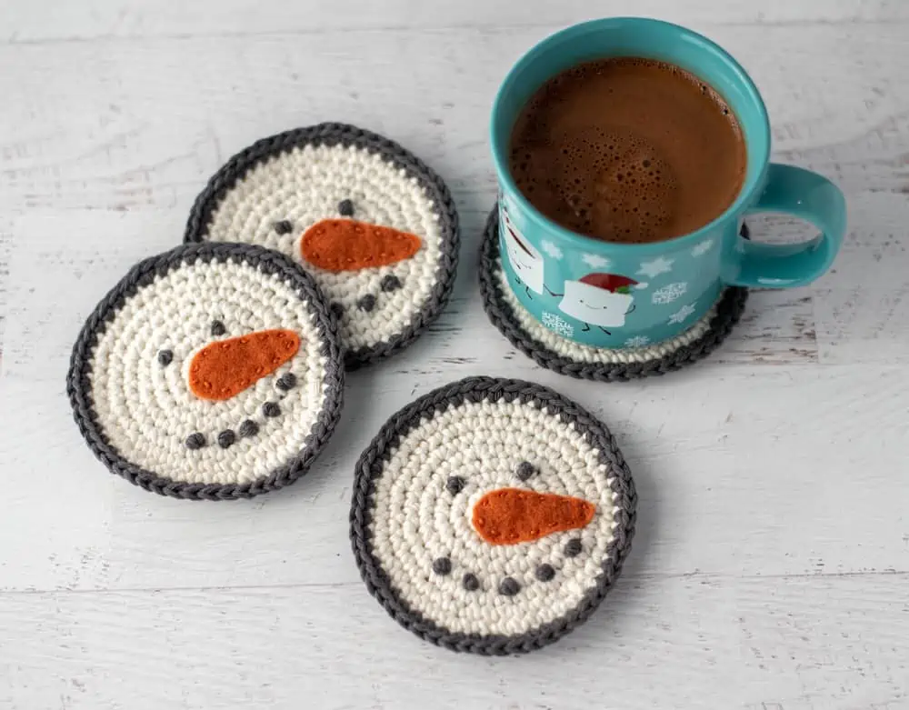 cream and gray crochet snowman coasters with orange carrot nose with cup of hot cocoa