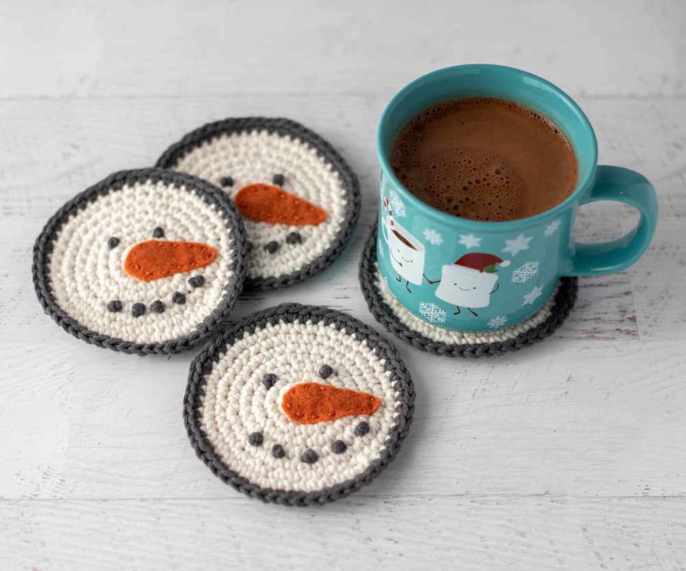 cream and gray crochet snowman coasters with orange carrot nose with cup of hot cocoa