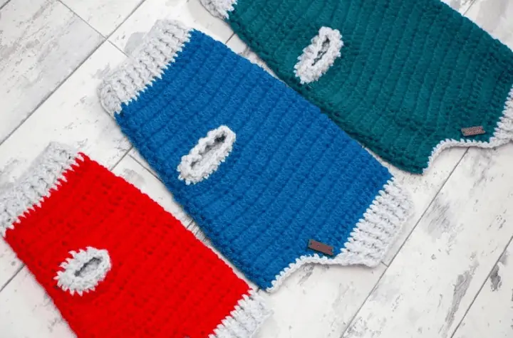 Three crochet dog sweaters, on is red with grey accents, one is blue with grey accents, and one is green with grey accents.