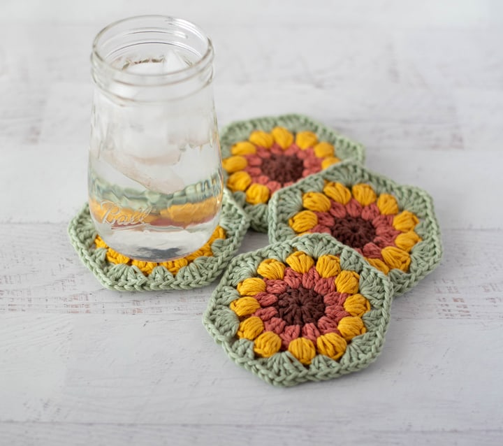 4 crochet sunflower coasters in brown, orange, yellow and green yarn with glass of ice water