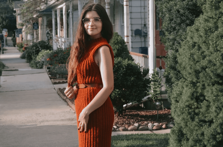 Long crochet sleeveless dress created using orange yarn and sinched at the waist with a brown belt.