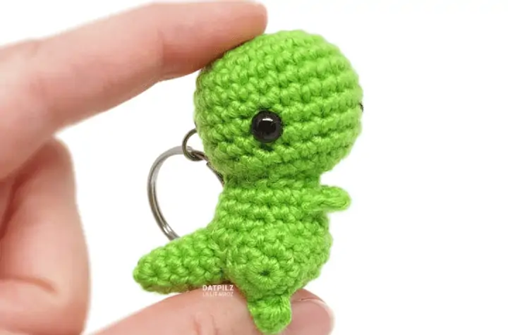 Green crochet T-Rex being held between fingertips.