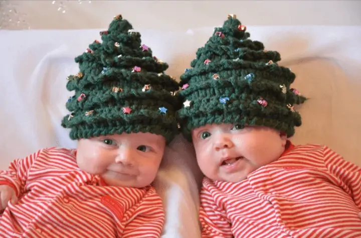 Two babies wearing crochet christmas tree hats with little stars on them.