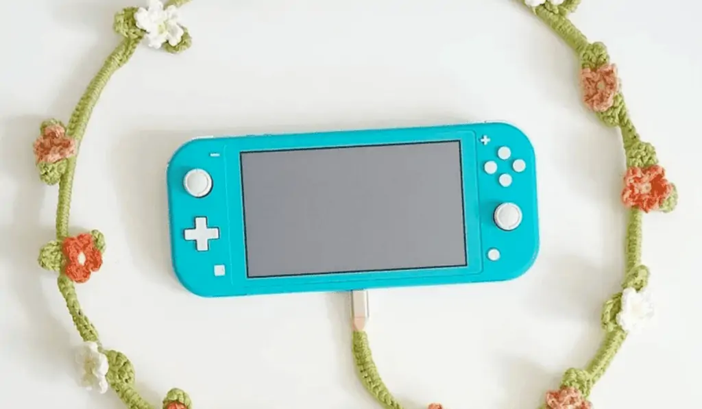 A crochet phone charger cover with little flowers around it being circled around a blue switch lite.