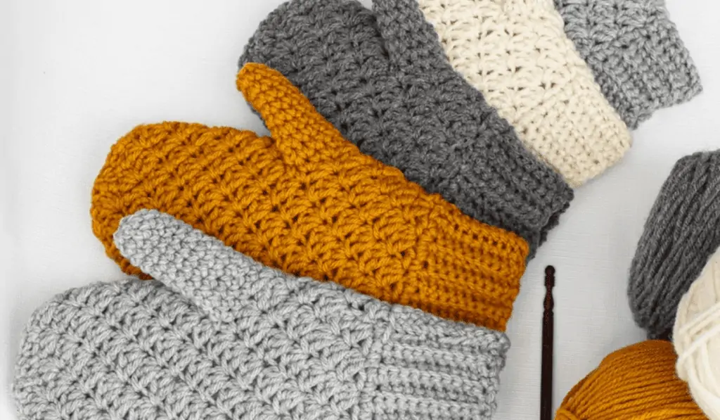 Crochet Mittons in different sizes, on light grey, one orange, one dark grey, one white, and one medium grey.