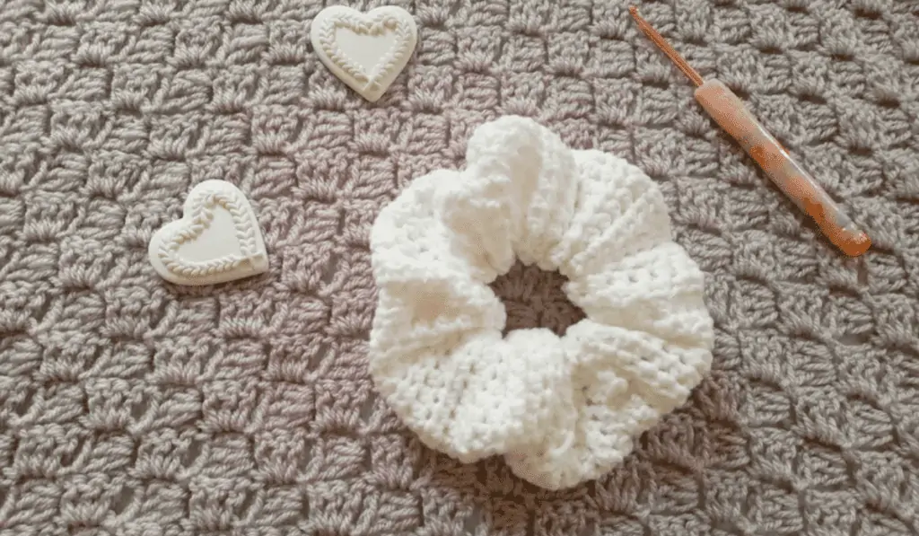 A white crochet scrunchie with a crochet hook sitting on top of a beige blanket.