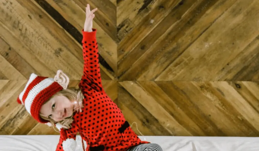 A little girl wearing a crochet elf hat with ears on the side with an arm sticking straight up in the air.