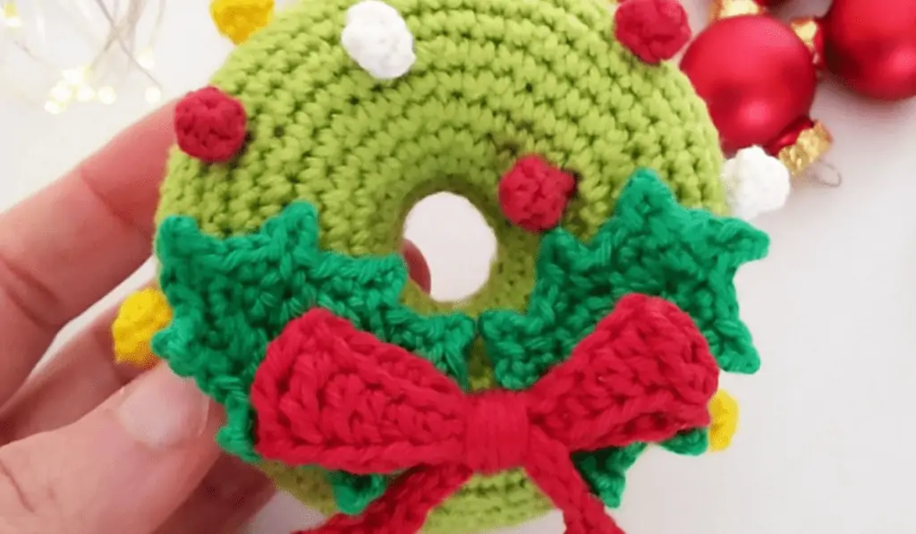 Someone holding a green crochet wreath that's stuffed like an amigurumi and features a little red bow.