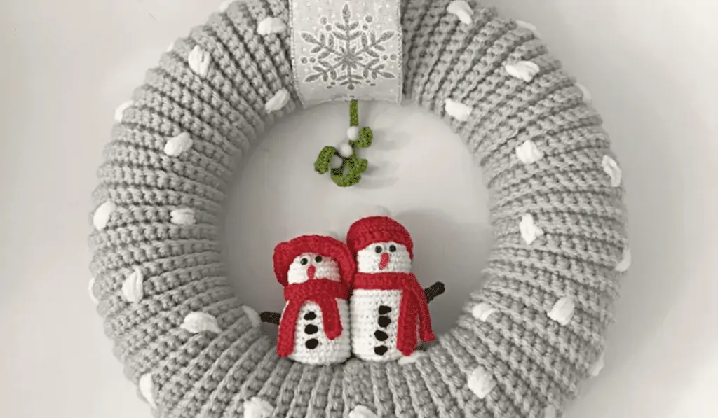 A grey crochet wreath with little white dots to simulate snow and two little snowmen under mistletoe in the center of this wreath.