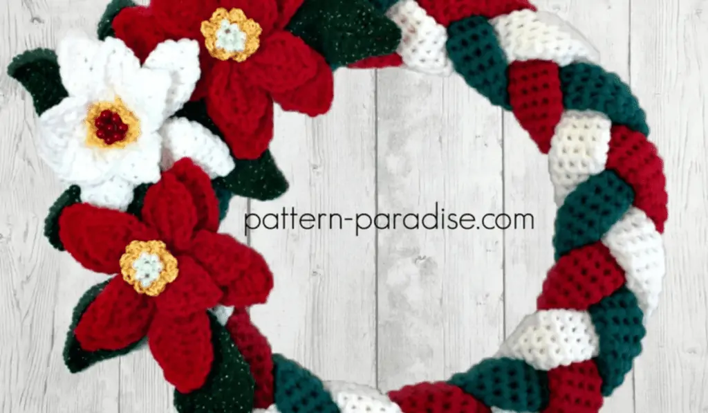 A crochet wreath with braided yarn strands in red, white, and green with three crocheted flowers in red and white.