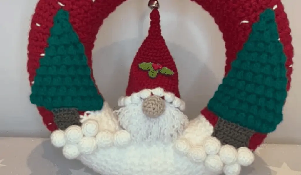 A red crochet wreath with two pine trees on either side and a gnome in the center. This gnome has a holly on it's hat and is standing on white yarn that looks like snow.