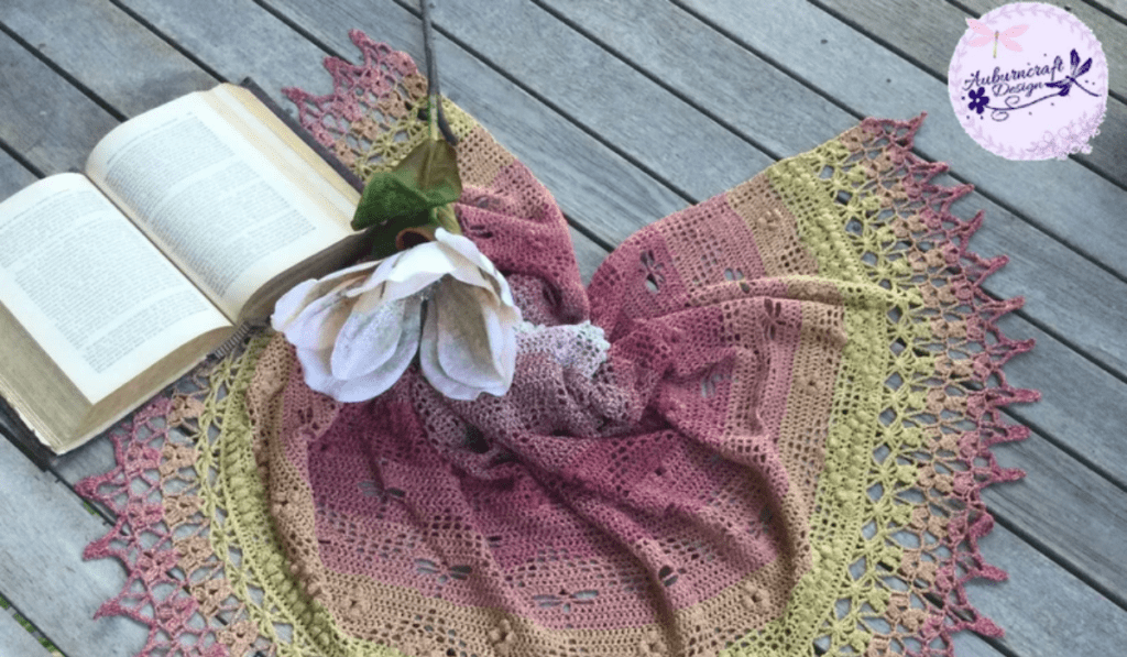 A multicolored shawl that's scrunched in the middle with a book off to the side and a flower laying over the top of it.