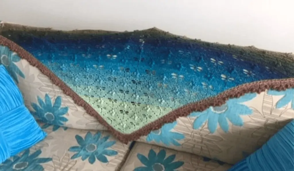 An ombre blue shawl with little dragonfly cut outs within the shawl drapped over the back of a couch.