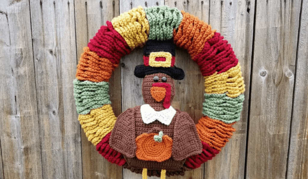 a crochet turkey wearing a pilgrim hat and holding a pumpkin on a wreath mad eout or red, orange, green, and yellow yarn.
