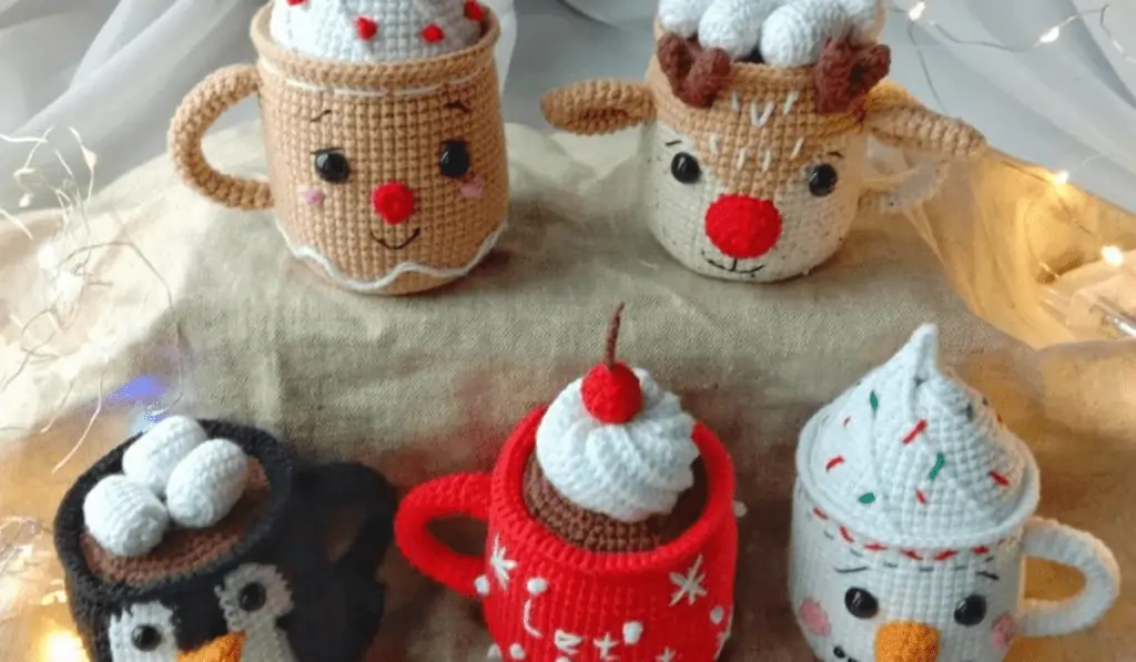 Five amigurumi mugs with crochet hot chocolate inside that look like different creatures, including a gingerbreadman, a reindeer, a penguin, a snowman, and then a mug that is red with snow falling on it.