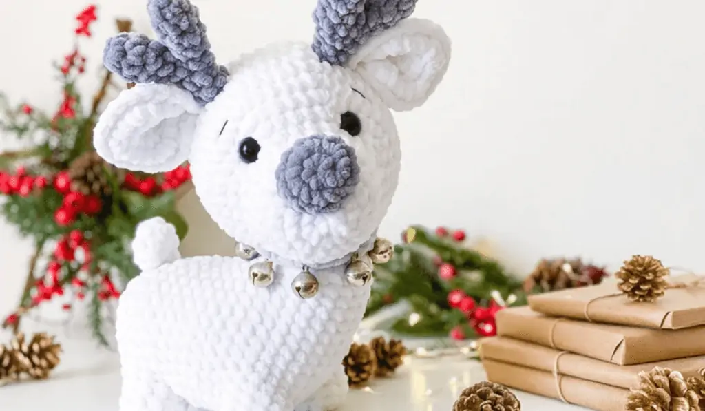 An all-white crochet reindeer with blue antlers and nose wearing a blue color with a lot of jingle bells around it.