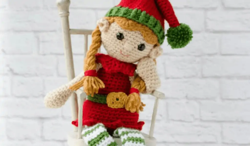 Crochet elf with blonde braided pigtails sititng in a wooden chair.