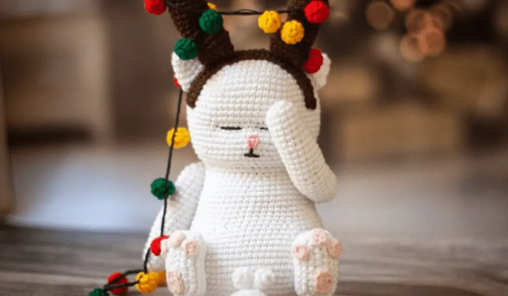 A white amigurumi cat face palming while wearing reindeer antlers with Christmas lights in them.