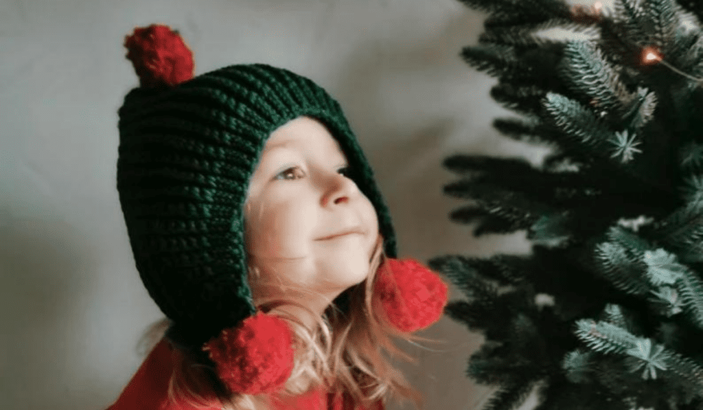 A little girl looking at a tree with a crochet green hood and red balls on the bottom.