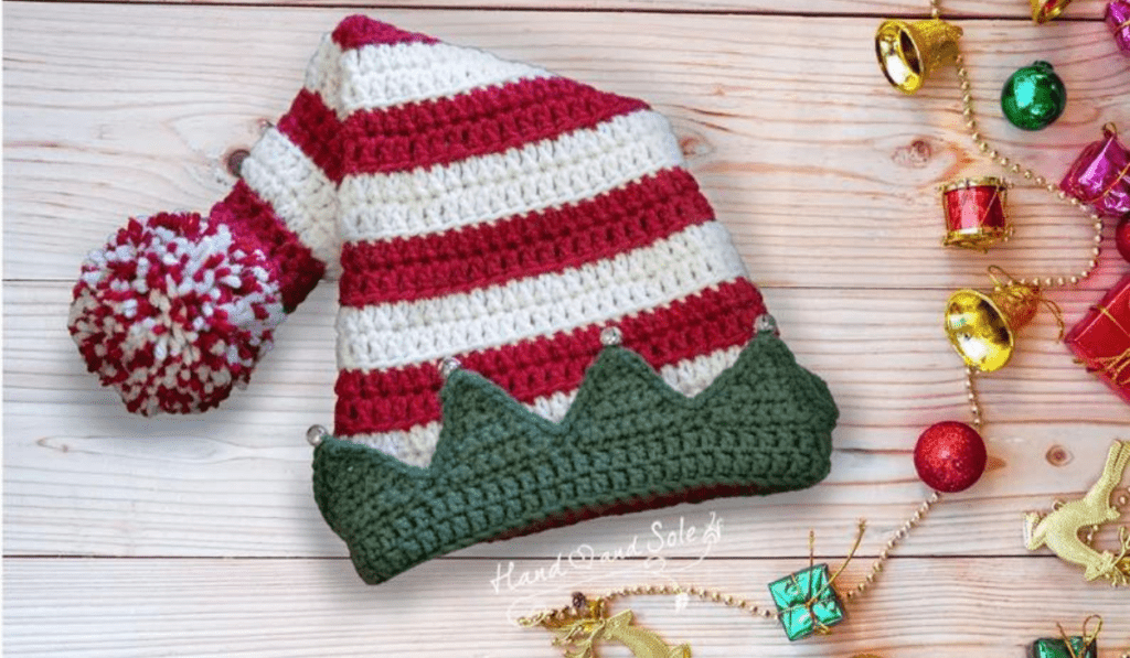 A red and white striped crochet elf hat with a green brim with triangle peaks with jingle bells on top.