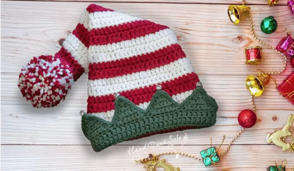 A red and white striped crochet elf hat with a green brim with triangle peaks with jingle bells on top.