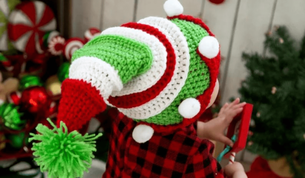 A little kid wearing a crochet elf hat made out of green, white, and red yarn with little white yarn balls on red triangle peaks along the brim.