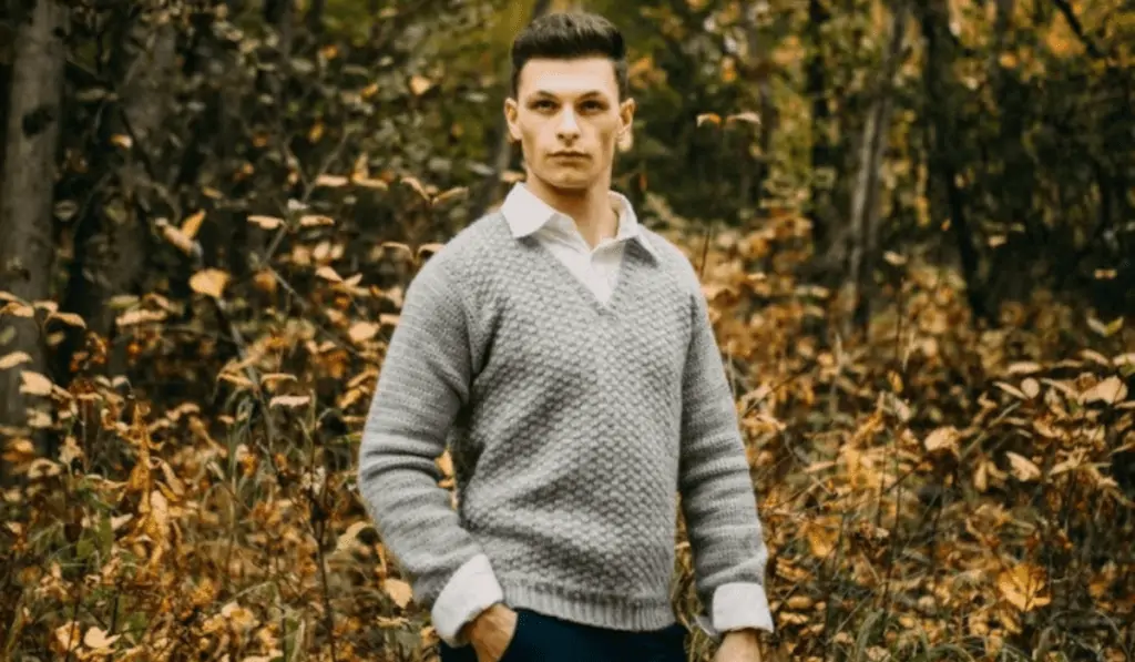 A man wearing a grey v-neck sweater with trees in the background.