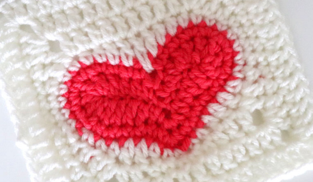 A white crochet granny square with a red heart in the middle.