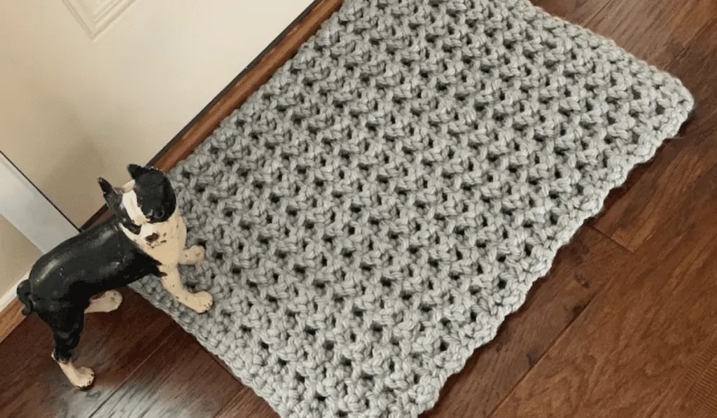 A grey crochet rug with a statue of a boston terrier next to it.