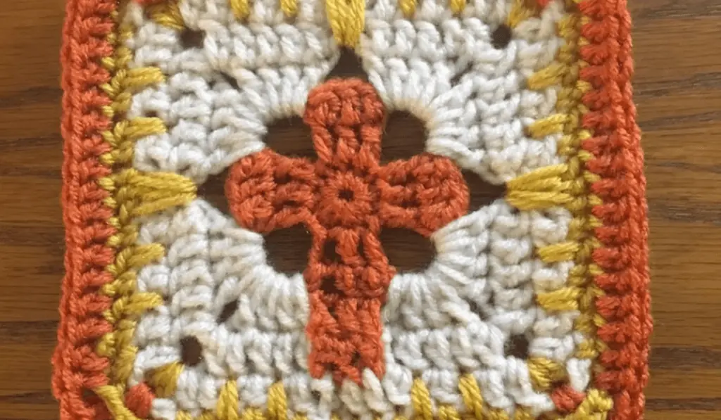A crochet cross granny square in orange, gold, and white yarn.