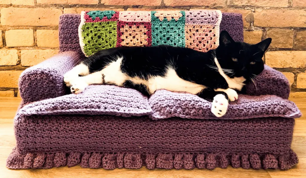 A purple crochet sofa cat bed with a small afghan on the back.