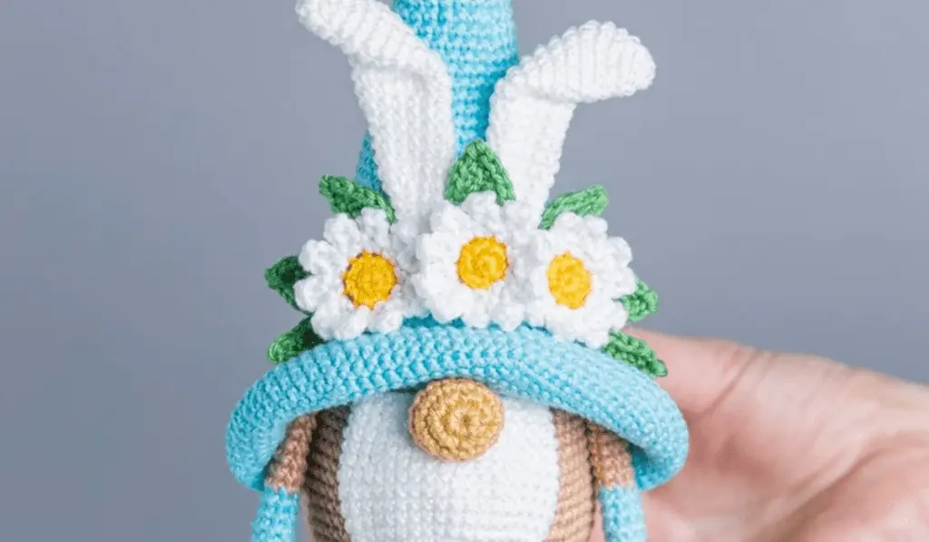 A crochet gnome with a blue hat that has bunny ears and white flowers on it.