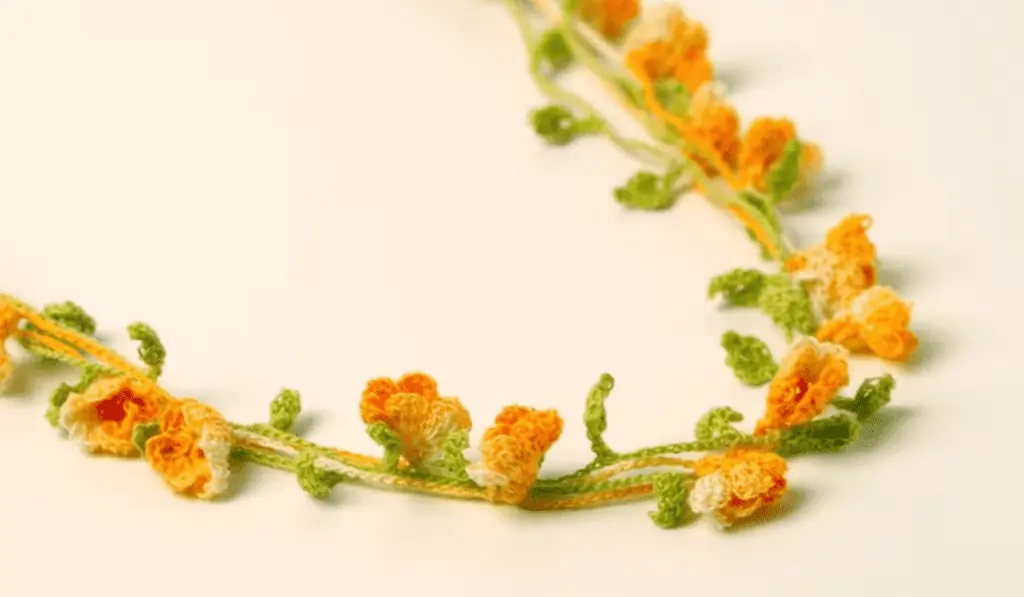 A crochet necklace with orange crochet flowers and a green vine to make up the chain of the necklace