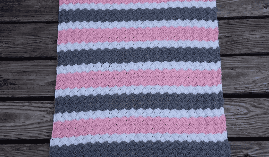 A bold color-blocked crochet blanket with stripes of pink, white, and grey yarn.