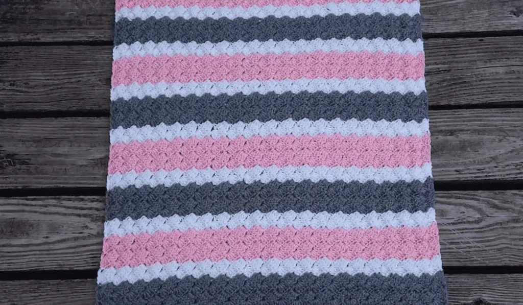A bold color-blocked crochet blanket with stripes of pink, white, and grey yarn.