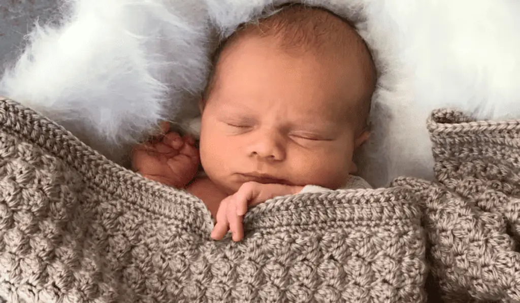 A baby tucked in with a beige crochet baby blanket